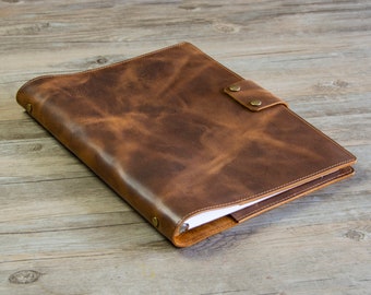 May include: A brown leather bound journal with a strap closure and brass accents. The journal is open to reveal blank pages.