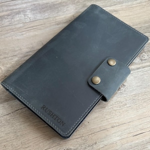 May include: A gray leather journal with the name "RUSHTON" embossed on the cover. The journal has a flap closure with two brass-colored snap closures. The journal is lying on a wooden surface.