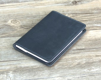 May include: A black leather notebook with white stitching. The notebook is closed and lying on a wooden surface.