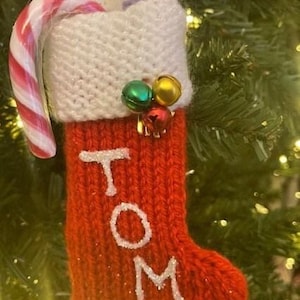 Puede incluir: Un calcetín navideño rojo tejido con el nombre "TOM" en purpurina blanca. El calcetín está decorado con un bastón de caramelo, pequeñas campanas y un puño blanco. Se ve un billete de veinte libras en el interior.