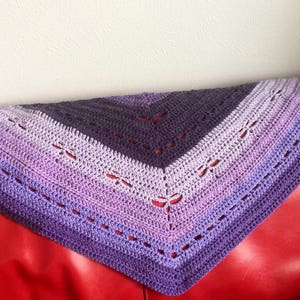 Puede incluir: Un chal de ganchillo morado y lavanda con un patrón de encaje. El chal está drapeado sobre un sofá rojo.