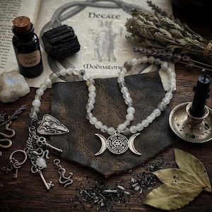 May include: A beaded necklace featuring a silver-toned triple moon pendant with a central labyrinth design. The necklace includes a cluster of charms, including a skull, keys, and a snake, attached to a dark leather-like piece on a wooden surface.