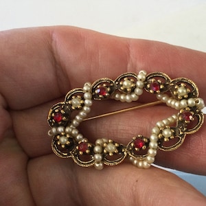 May include: A gold-toned brooch with a delicate filigree design. The brooch is decorated with small white pearls and red rhinestones.