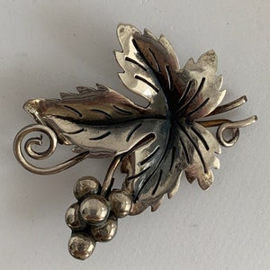 May include: A silver brooch in the shape of a grapevine leaf with a cluster of grapes. The leaf is detailed with veins and the grapes are round and smooth.