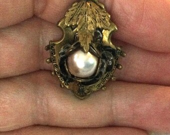 May include: A gold-toned brooch with a pearl in the center, surrounded by a black and gold design. The brooch is shaped like a shield and has a leaf design on top.