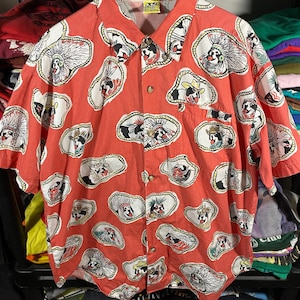 May include: A short-sleeved button-down shirt in a coral color, featuring a repeating pattern of cartoon dogs in Native American headdresses and cowboy hats. The shirt has a chest pocket and a collar.