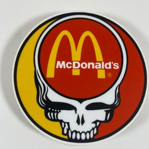May include: A round sticker with a yellow and red background. The red part features the McDonald's logo with the text "McDonald's" and a registered trademark symbol. The yellow part features a black and white skull with a single eye and a smile.