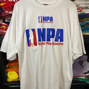 May include: White t-shirt with the text "NPA National Pimp Association" in blue and red. The shirt features a graphic of a person in a hat and a cane. The shirt is hanging on a wooden hanger.
