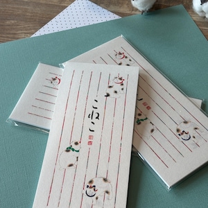 Può includere: Tre buste rettangolari in carta color bianco sporco con linee verticali rosse e verdi e illustrazioni di gatti calico con collari. Il testo giapponese "こねこ" è stampato verticalmente in inchiostro nero al centro.