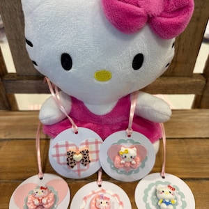 Puede incluir: Una muñeca de peluche blanca de Hello Kitty con un lazo y un vestido rosa, sosteniendo varias etiquetas redondas. Las etiquetas presentan diseños de Hello Kitty con lazos y corazones. Las etiquetas están unidas con cintas rosas.