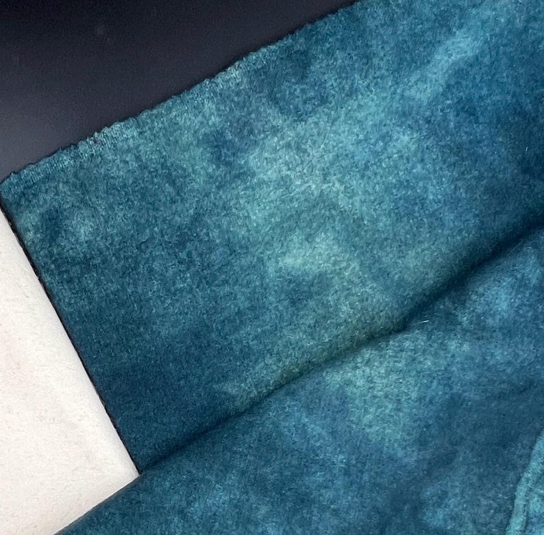 May include: Close-up of a teal-colored fabric with a soft, velvety texture. The fabric has a mottled appearance with darker and lighter shades of teal, creating a watercolor effect. The fabric is folded, revealing its thickness and drape.