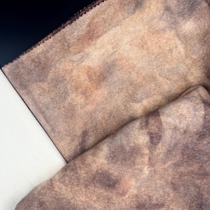 Peut inclure: Un morceau de tissu présentant un motif marbré de style tie-dye dans des tons de brun, de beige et de violet atténué. Le textile a un aspect doux et légèrement texturé avec un bord plié visible et un ourlet cousu brun foncé sur le haut.