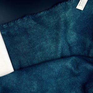 May include: Close-up of a textured teal fabric with a dark blue leather-like material and white fabric. The teal fabric has a slightly frayed edge. A white tag with text is visible in the upper right corner.
