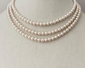 May include: A multi-strand necklace featuring three distinct layers of uniform, cream-colored spherical pearls. Each strand is composed of small, smooth, round beads arranged in a classic, cascading circular design that rests evenly around the neck.