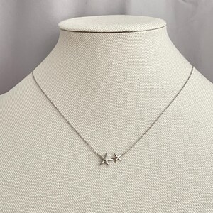 May include: A delicate silver-toned necklace featuring two small starfish charms positioned side-by-side at the center of a thin, fine link chain. The metallic starfish pendants have a smooth, polished finish and are attached directly to the chain.