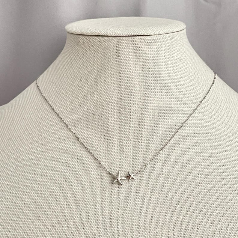 May include: A delicate silver-toned necklace featuring two small starfish charms positioned side-by-side at the center of a thin, fine link chain. The metallic starfish pendants have a smooth, polished finish and are attached directly to the chain.
