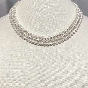 May include: A three-strand pearl necklace displayed on a mannequin. The necklace features small, round, off-white pearls. The strands are closely set, creating a full, elegant look. The necklace is draped in a gentle curve.