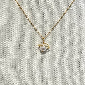 Può includere: Una collana con catena dorata e un ciondolo di perla. Il ciondolo è circondato da un sottile anello dorato, che ricorda un pianeta con un anello.
