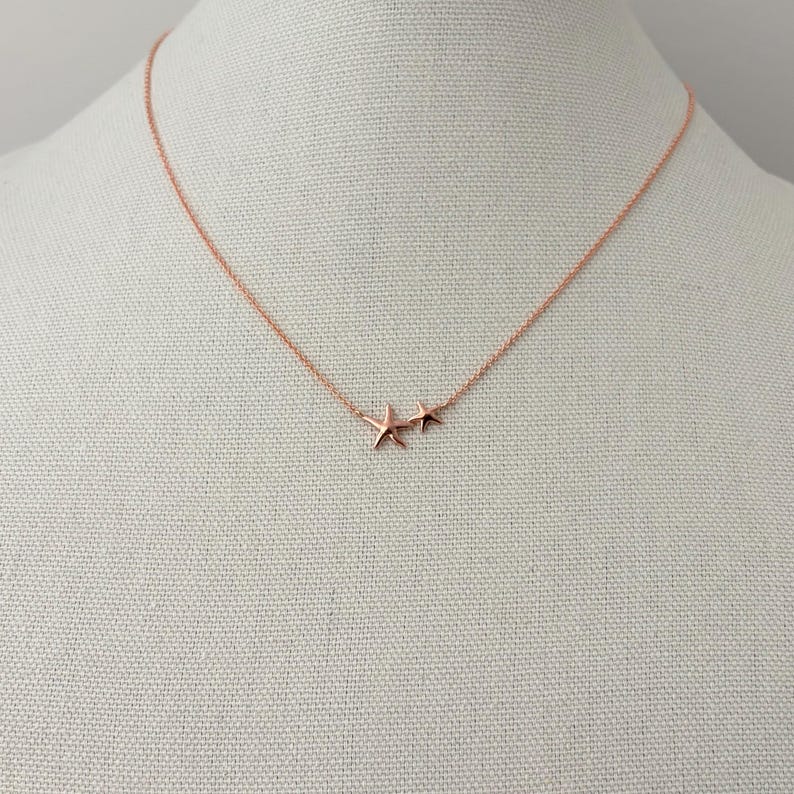 May include: A rose gold necklace featuring a delicate chain and two small starfish charms of different sizes positioned together. The metal has a smooth, polished finish, and the starfish pendants are attached directly to the thin, circular link chain.