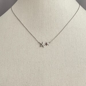 May include: A silver-toned necklace featuring a delicate chain and two small, five-pointed starfish charms positioned off-center. The metal has a smooth, polished finish, and the two starfish are of slightly different sizes, resting side by side.