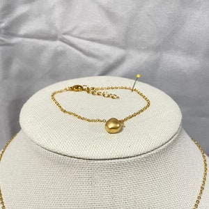 Puede incluir: Un brazalete de cadena dorada con un pequeño dije redondo dorado. El brazalete está en un expositor blanco.
