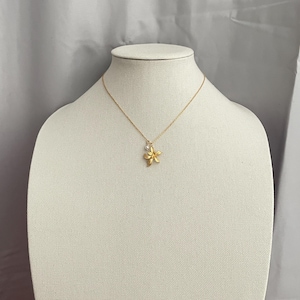 May include: A gold-colored necklace with a flower pendant and a small, round, clear gem. The necklace is displayed on a white mannequin. The flower pendant has five petals and is slightly textured.