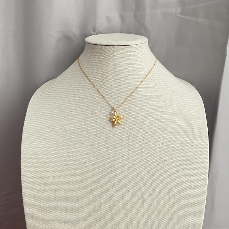 May include: A gold-colored necklace with a flower pendant and a small, round, clear gem. The necklace is displayed on a white mannequin. The flower pendant has five petals and is slightly textured.