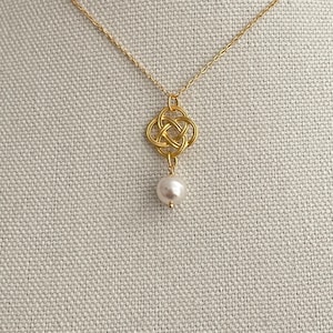 Op de afbeelding: Een goudkleurige ketting met een Keltische knoop hanger en een witte parel. De hanger is een gedetailleerd knoopontwerp en de parel hangt eronder. De ketting is een delicaat sieraad, geschikt voor dagelijks gebruik of speciale gelegenheden.