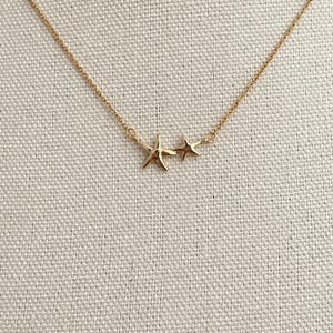 May include: A gold-toned necklace featuring a delicate chain and two connected star-shaped charms. The larger star is positioned on the left, while a smaller, simpler star sits to its right, creating a balanced and symmetrical design element.