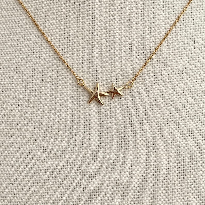 May include: A gold-toned necklace featuring a delicate chain and two connected star-shaped charms. The larger star is positioned on the left, while a smaller, simpler star sits to its right, creating a balanced and symmetrical design element.