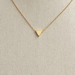 Peut inclure: Un collier doré délicat avec un petit pendentif triangulaire. Le pendentif est suspendu à une fine chaîne, créant un design minimaliste. Le collier est présenté sur un fond de tissu neutre.