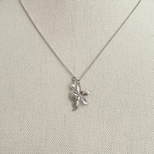 May include: A silver-toned necklace featuring a flower pendant with five petals and a small pearl charm. The pendant is detailed with etched lines, and the chain is delicate. The necklace is displayed on a neutral fabric background.