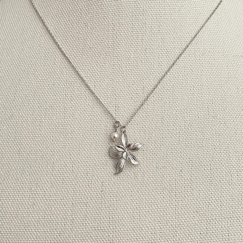 May include: A silver-toned necklace featuring a flower pendant with five petals and a small pearl charm. The pendant is detailed with etched lines, and the chain is delicate. The necklace is displayed on a neutral fabric background.