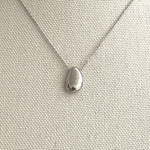 May include: A silver-colored necklace with a small, smooth, teardrop-shaped pendant. The pendant hangs from a delicate chain. The background is a neutral, textured fabric.