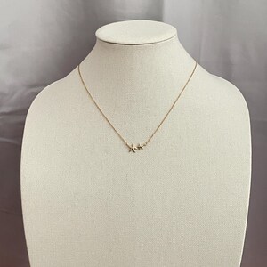 May include: A gold-toned necklace featuring a delicate chain and a pendant consisting of two small, five-pointed starfish charms positioned side-by-side. The metal has a polished finish, and the necklace is displayed on a neutral-colored form.