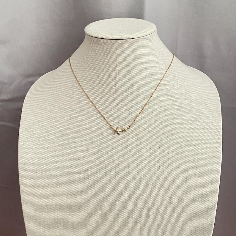 May include: A gold-toned necklace featuring a delicate chain and a pendant consisting of two small, five-pointed starfish charms positioned side-by-side. The metal has a polished finish, and the necklace is displayed on a neutral-colored form.