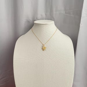May include: A gold-colored necklace with a flower pendant and a small white pearl. The necklace is displayed on a white mannequin bust against a gray backdrop. The flower pendant has detailed petals.