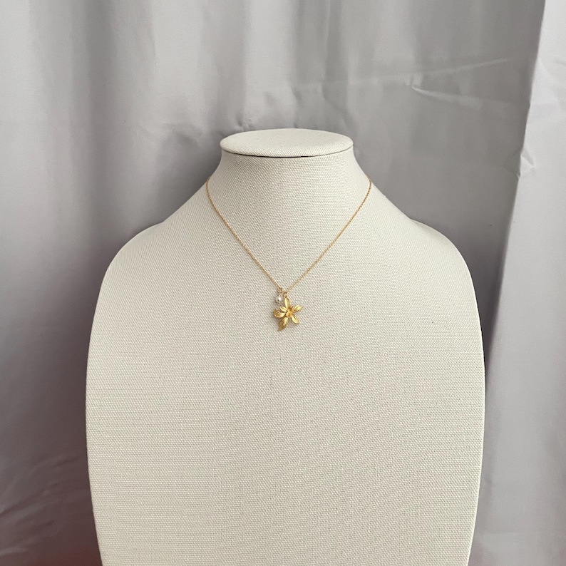 May include: A gold-colored necklace with a flower pendant and a small white pearl. The necklace is displayed on a white mannequin bust against a gray backdrop. The flower pendant has detailed petals.