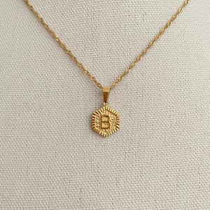 May include: A gold chain necklace with a hexagonal pendant featuring the letter "B" in a textured design.