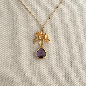 May include: A gold-colored necklace featuring a lotus flower pendant with a purple gemstone. The pendant has a detailed lotus flower design above a teardrop-shaped, dark purple gemstone. The necklace is displayed on a textured, light-colored surface.