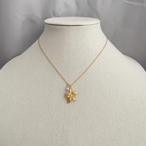 May include: A gold-colored necklace with a flower pendant and a small pearl. The flower pendant has five petals and is suspended from a delicate gold chain. The necklace is displayed on a mannequin.