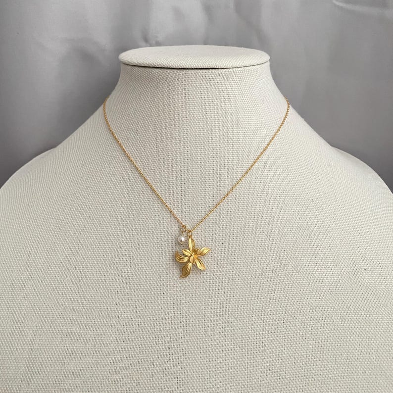 May include: A gold-colored necklace with a flower pendant and a small pearl. The flower pendant has five petals and is suspended from a delicate gold chain. The necklace is displayed on a mannequin.