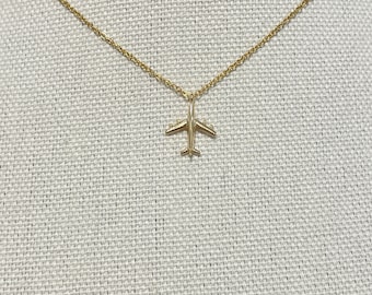 May include: A gold-colored airplane pendant necklace. The pendant is a detailed miniature of an airplane. The necklace chain is delicate and also gold-colored. The necklace is displayed on a textured, off-white surface.