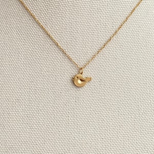 May include: A gold-toned necklace featuring a delicate chain and a small, smooth bird-shaped pendant. The pendant is minimalist in form, depicting a stylized bird in profile with a tiny dot for an eye, suspended from a thin, polished metal chain.