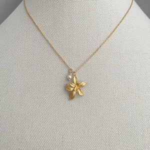 May include: A gold-colored necklace with a delicate chain and a flower pendant. The pendant has a detailed floral design and a small pearl charm. The necklace is displayed on a neutral fabric background.