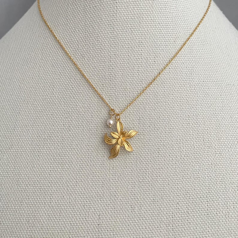 May include: A gold-colored necklace with a delicate chain and a flower pendant. The pendant has a detailed floral design and a small pearl charm. The necklace is displayed on a neutral fabric background.