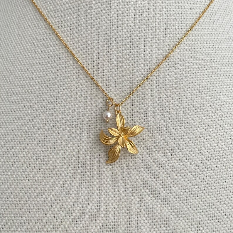 May include: A gold-colored necklace featuring a flower pendant and a small pearl charm. The flower pendant has detailed petals. The necklace is displayed on a neutral-colored fabric.