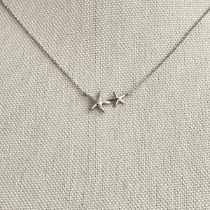 May include: A silver-toned necklace featuring two connected starfish charms of different sizes. The pendant is suspended from a delicate, thin metal chain with a polished finish, creating a simple and clean look for this piece of jewelry.