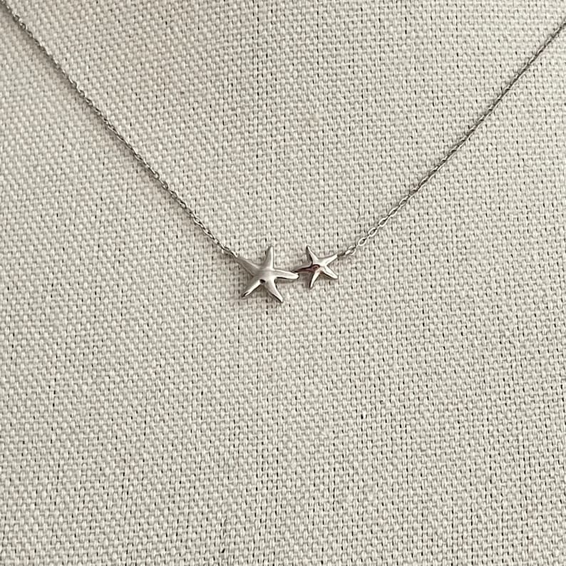 May include: A silver-toned necklace featuring two connected starfish charms of different sizes. The pendant is suspended from a delicate, thin metal chain with a polished finish, creating a simple and clean look for this piece of jewelry.
