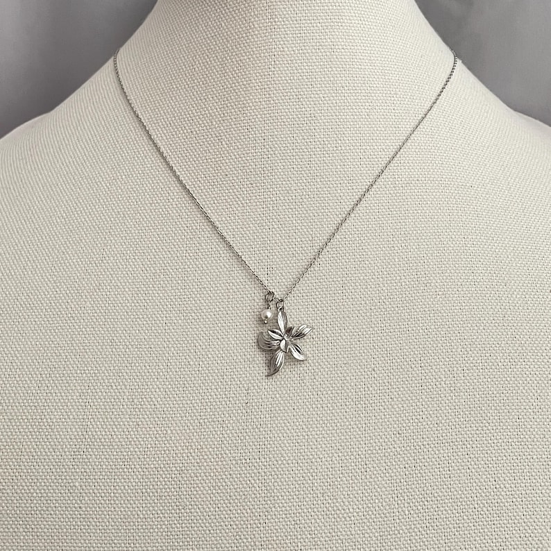 May include: A silver-toned necklace featuring a floral pendant with a small pearl accent. The pendant is a detailed, five-petal flower design. The necklace is displayed on a neutral-colored mannequin.